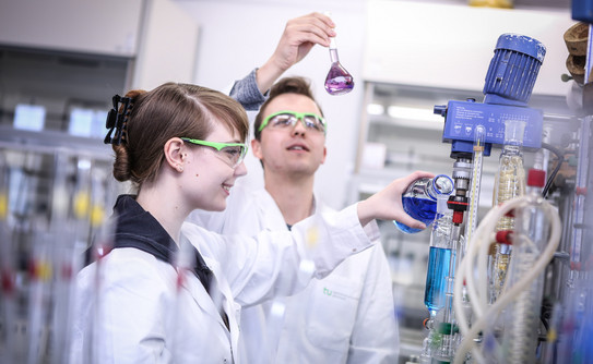 Auszubildende im Chemielabor_01 Zwei Auszubildende in einem chemischen Labor mit weißen Kitteln, einer gießt blaue Flüssigkeit in ein Destilliergerät.