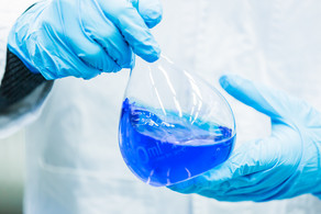 Detailaufnahme Messkolben Zwei Hände in blauen Gummihandschuhen halten einen Messkolben, der mit blauer Flüssigkeit gefüllt ist.